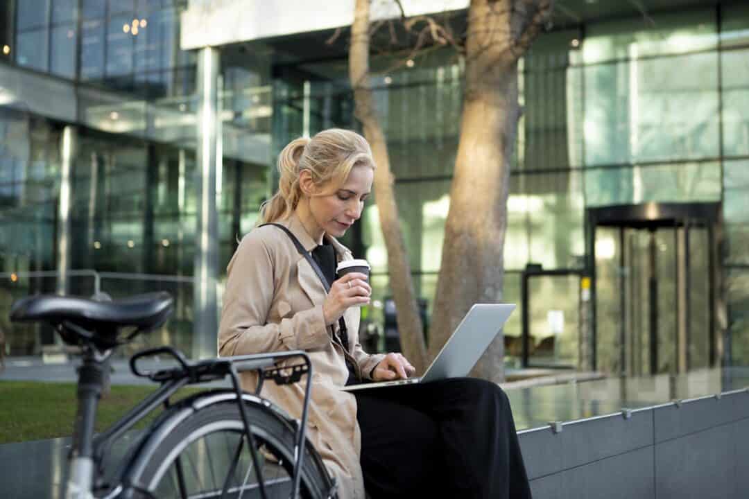 En kvinde sidder udendørs og drikker kaffe og bruger en bærbar computer.